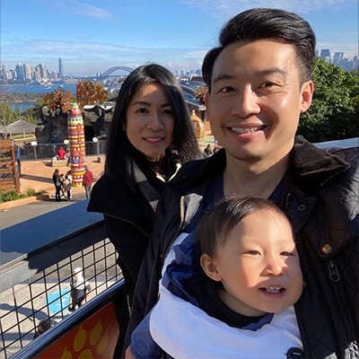 A family of three smiling for a selfie with a city skyline featuring a bridge and a large tower in the background on a sunny day.