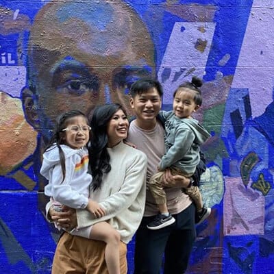 A family of four smiling in front of a vibrant mural, with the parents holding their two young daughters.