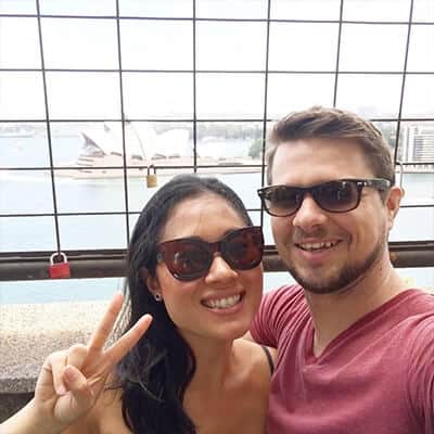 A smiling couple in sunglasses takes a selfie with a wire fence and harbor background, woman making a peace sign.