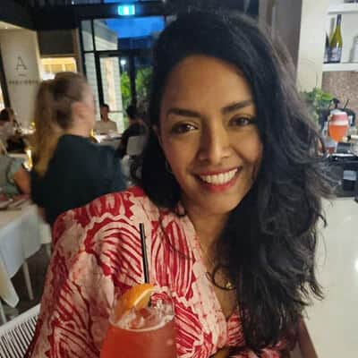 A woman smiling at a restaurant table, holding a cocktail, wearing a red and white floral dress.