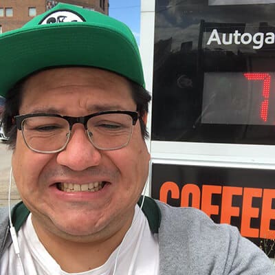 Man in a green cap smiling with a squint beside a coffee shop sign showing the time as 7 o'clock.