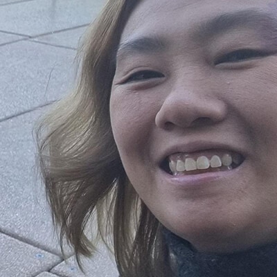 Close-up of a smiling person with shoulder-length light brown hair and a black turtleneck, outdoors.