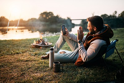 Man sitting in a camping chair by a river at sunset, using a smartphone, with a campfire in the background.