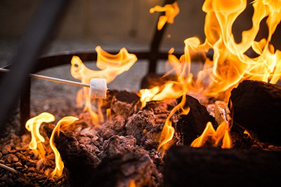 A marshmallow on a skewer being roasted over a vibrant campfire, with flames engulfing logs and embers glowing beneath.