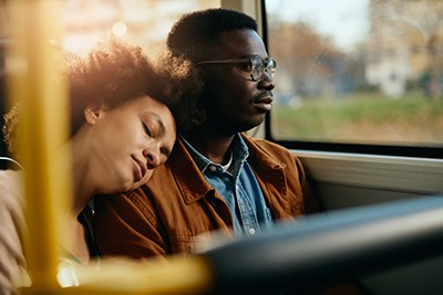 A woman rests her head on a man's shoulder as they ride a bus, with sunlight streaming through the window beside them.